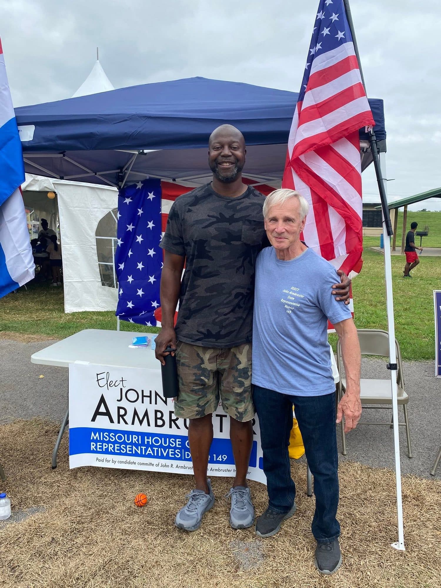 John Armbruster, in a campaign T-shirt, stands with a supporter at an outdoor community event beneath the campaign's tent.